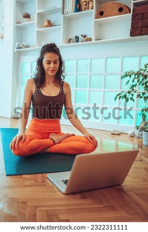 Wide shot of a caucasian young girl hosting an online meeting on her laptop while sitting in a lotus pose on her yoga mat in a cozy studio.Looking down and smiling. Remote work concept. Copy space.