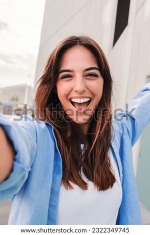 Vertical portrait of a happy young caucasian student lady looking at camera and taking a selfie having fun, standing outside. Laughing woman shooting a photo for social media at the university campus