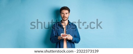Image of smiling young man celebrating birthday, holding b-day cake with lit candle and looking at camera, standing over blue background.
