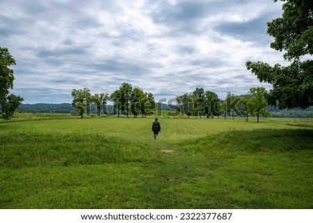 A young man walks toward the Native American Hopewell Culture prehistoric Seip Earthworks burial mound in Ohio. Ancient large long mound. Grass is neatly trimmed with trees dramatic sky. Copy space