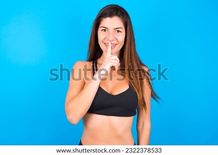 Smiling Young beautiful brunette woman wearing sportswear makes shush gesture, holds fore finger over lips hides secret. Be mute, please.