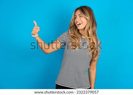 Young beautiful woman wearing striped t-shirt Looking proud, smiling doing thumbs up gesture to the side. Good job!