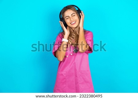 Pleased young beautiful doctor woman standing over blue studio background enjoys listening pleasant melody keeps hands on stereo headphones closes eyes. Spending free time with music Royalty-Free Stock Photo #2322380495