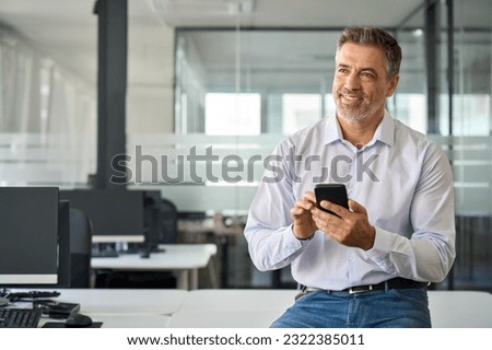 Happy mid aged business man using phone in office. Smiling mature confident businessman executive manager holding smartphone working on cell phone sitting at office desk looking away and thinking. Royalty-Free Stock Photo #2322385011