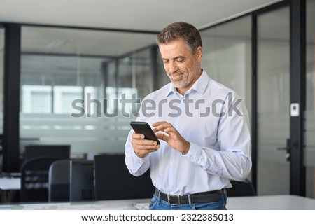Smiling older business man executive standing in office using mobile phone. Happy mid aged professional manager holding cell working on smartphone checking financial transactions on cellphone tech.