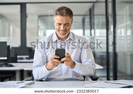 Happy older mid aged business man ceo executive, mature businessman manager sitting in modern office holding smartphone using mobile cell phone managing digital tech transactions on cellphone at work.