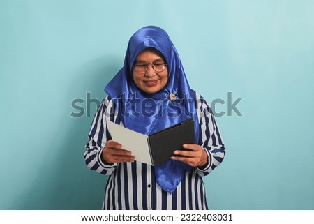 Amazed Asian middle-aged woman, wearing a blue hijab, eyeglasses, and a striped shirt, holds an open book with a surprised expression while standing against a blue background.