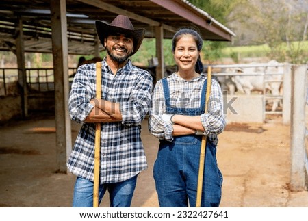 Portrait of Asian man and woman farmers carry broom and stand with arm crossed also look at camera with smiling in front of stable of cow look relax after clean the area.