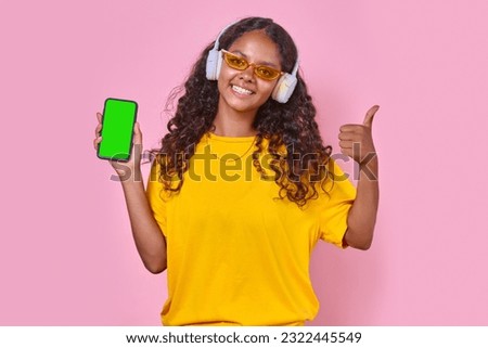 Young attractive positive Indian woman teen in wireless headphones holds mobile phone and shows thumbs up to praise new album of popular singer or music group stands in pink studio. Green screen