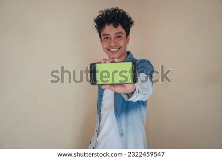 Young curly man wear denim clothes hold smart phone with green screen. Smile, cheers and happy expression. The photo is suitable to use for man expression advertising and fashion lifestyle.