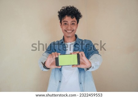 Young curly man wear denim clothes hold smart phone with green screen. Smile, cheers and happy expression. The photo is suitable to use for man expression advertising and fashion lifestyle.