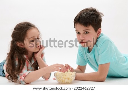 Beautiful primary school children, lovely little kid girl in pink blue plaid shirt and her older brother, pre teen charming boy in t-shirt, lying on belly, eating popcorn on isolated white background.