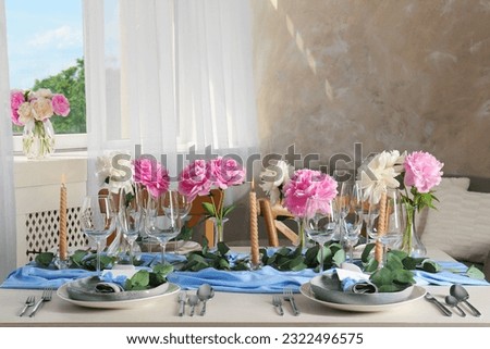 Beautiful table setting. Plates with greeting cards, napkins and branches near glasses, peonies, burning candles and cutlery on table in room