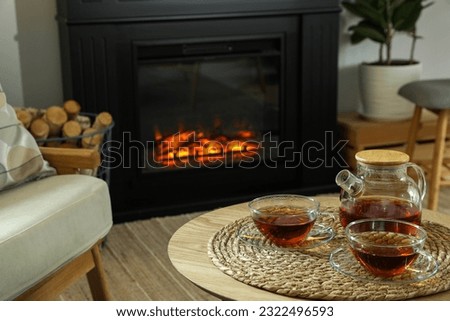 Teapot and cups of drink on coffee table near stylish fireplace in cosy living room. Interior design
