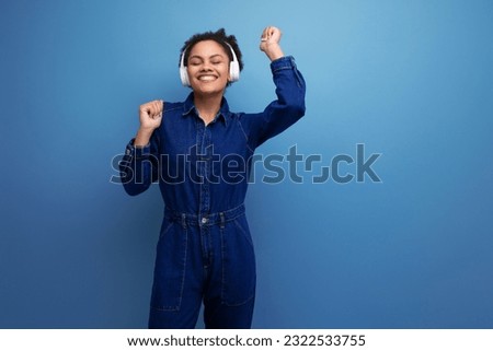 young joyful brunette latin woman dressed in blue denim overalls listens to music in wireless headphones on a blue background Royalty-Free Stock Photo #2322533755
