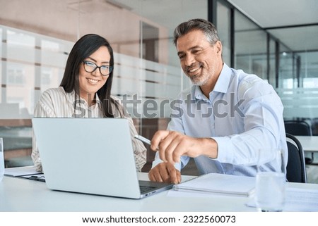 Mid aged Latin male manager mentor teaching young Asian female worker looking at laptop discussing corporate strategy in teamwork, working on computer in office at international team meeting. Royalty-Free Stock Photo #2322560073