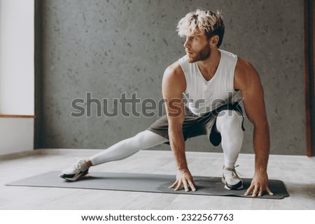Full body young strong sporty athletic sportsman man wearing white tank shirt black shorts doing stretch exercise for legs sit at floor look aside warm up training indoor at gym Workout sport concept Royalty-Free Stock Photo #2322567763