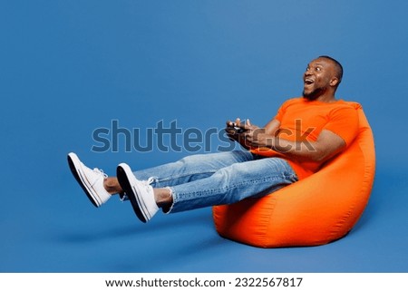 Full body young man of African American ethnicity wear orange t-shirt sit in bag chair hold in hand play pc game with joystick console isolated on plain dark royal navy blue background studio portrait