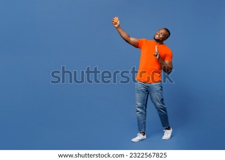 Full body young man of African American ethnicity he wear orange t-shirt doing selfie shot on mobile cell phone post photo on social network isolated on plain dark royal navy blue background studio
