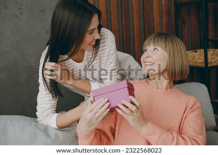 Two smiing happy adult women mature mom young kid wearing casual clothes give pink present box sit on gray sofa couch stay at home flat rest relax spend free spare time in living room. Family concept