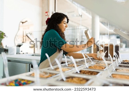 Young woman worker smiling while preparing delicious frozen yogurt with toppings while working behind the bar counter at the shop Royalty-Free Stock Photo #2322593473
