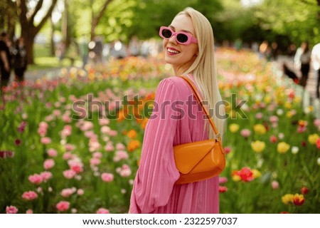 Happy smiling fashionable woman wearing trendy pink sunglasses, pleated dress, with orange faux patent leather baguette bag, posing among flowers. Copy, empty space for text Royalty-Free Stock Photo #2322597037
