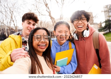 Group of multi-ethnic high school students taking a selfie outdoors at the university campus holding folders. Looking at camera picture of a diverse colleagues having fun. 