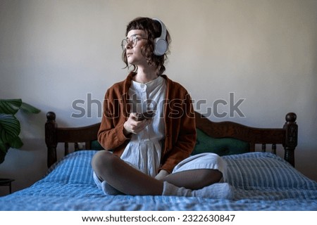Millennial woman listen to music at home, teenage girl sitting on bed in lotus pose using smartphone and headphones while relaxing in bedroom. Learning through listening, podcasting concept