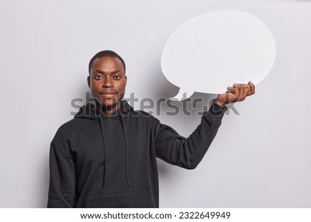 Waist up shot of dark skinned man holds empty blank speech bubble eagerly waiting to fill it with words that convey his thoughts or message wears casual black sweatshirt isolated over white background