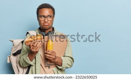 Photo of displeased dark skinned man wears spectacles and headphones around his neck eats hot dog drinks orange juice carries notepads and rucksack isolated over blue background copy space for ttext
