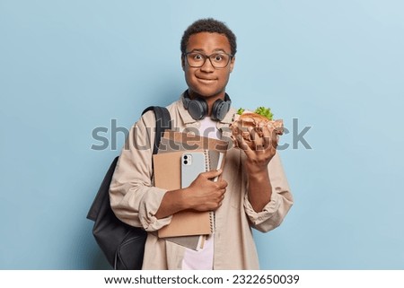 Education and lifestyle concept. Handsome dark skinned male student eats sandwich after day of studying holds spiral notepads carries rucksack dressed casually isolated over blue background. Royalty-Free Stock Photo #2322650039