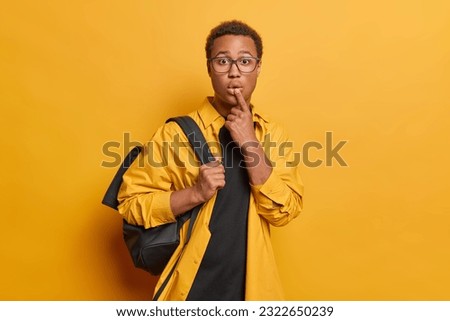 Surprised teenage guy goes to university wears shirt and spectacles carries rucksack keeps lips rounded reacts to something impressive isolated over vivid yellow background. Stunned male student
