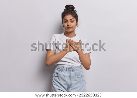 Tender Iranian girl pressing palms to her heart with sense of gratitude and admiration expressing heartfelt sentiments has serious expression being thankful wears casual t shirt jeans isolated
