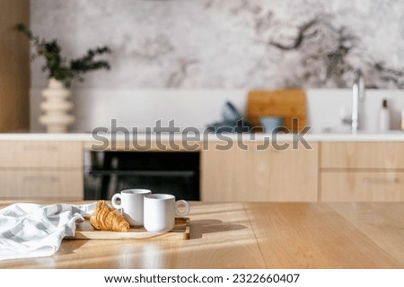 dish towel, fresh croissant and ceramic cups of tea on bamboo tray on wooden tabletop with sun light on kitchen background interior, breakfast concept Royalty-Free Stock Photo #2322660407