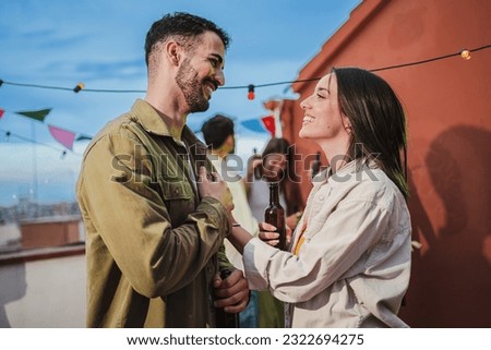 Smiling young couple flirting and having fun at a rooftop party with friends, drinking beer, enjoying and having a pleasant conversation. Handsome guy greeting his joyful girlfriend at celebration Royalty-Free Stock Photo #2322694275