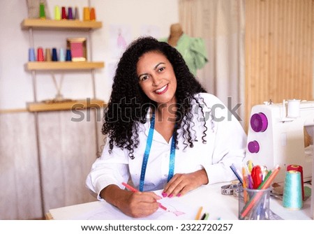 Fashion Design. Cheerful Hispanic Tailor Woman Sketching New Clothing Patterns Sitting At Sewing Machine In Dressmaking Showroom Indoor, Smiling At Camera. Successful Dressmaking Business