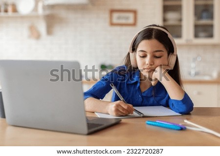 Boring Online Education. Bored Schooler Girl Learning At Laptop And Taking Notes Sitting With Earphones At Desk At Home. Tired School Kid Doing Difficult Homework Indoors