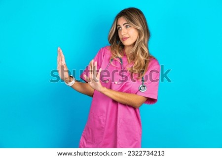 Displeased young beautiful doctor woman standing over blue studio background keeps hands towards empty space and asks not come closer sees something unpleasant