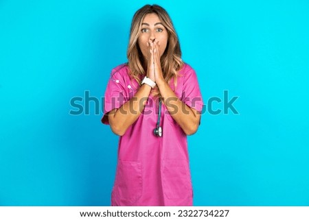 young beautiful doctor woman standing over blue studio background keeps hands on mouth, looks with eyes full of disbelief, being puzzled with amount of work