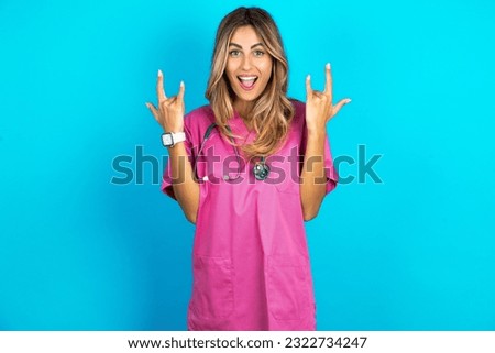 young beautiful doctor woman standing over blue studio background makes rock n roll sign looks self confident and cheerful enjoys cool music at party. Body language concept.