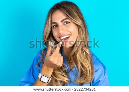 young beautiful doctor woman standing over blue studio background holding an invisible aligner ready to use it. Dental healthcare and confidence concept. Royalty-Free Stock Photo #2322734281