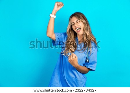 Profile photo of excited young beautiful doctor woman standing over blue studio background good mood raise fists screaming rejoicing overjoyed basketball sports fan supporter