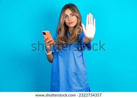 young beautiful doctor woman standing over blue studio background using and texting with smartphone with open hand doing stop sign with serious and confident expression, defense gesture
