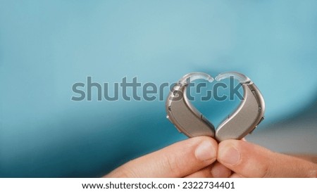 Hearing aids in hands making heart shape over blue background. Closeup of listening device for people with hearing disorder, disfunction. Technology that gives better sense of sound, speach Royalty-Free Stock Photo #2322734401