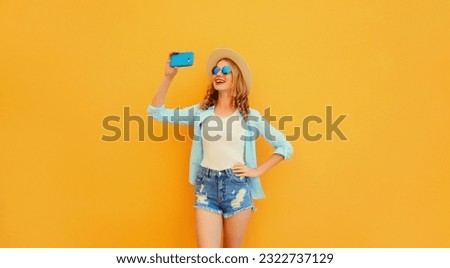 Portrait of happy smiling young woman taking selfie with smartphone wearing straw hat on yellow background