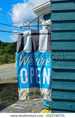 A blue, black, and white colored cloth flag hanging with the text We are open come on in. The banner advertisement is attached outside to the wall of a teal-green wooden cafe shop under blue sky.