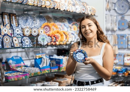 Enthusiastic young woman choosing decorative plate with colorful pattern in specialized souvenir shop