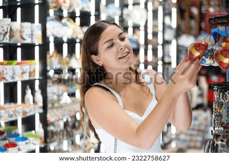 Happy interested young tourist girl looking for handmade crafted items on stands in souvenir shop .. Royalty-Free Stock Photo #2322758627