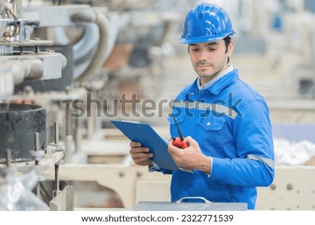 half body photo of a smart-looking American engineer holding a listnote, a walkie-talkie, looking down at machines, wearing uniforms, in a large plastics processing plant. There is a working machine