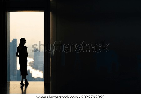 Silhouette Asian woman standing by the window at skyscraper building in metropolis and looking cityscape at summer sunset. Beautiful girl enjoy urban lifestyle activity in the city on holiday vacation Royalty-Free Stock Photo #2322792093